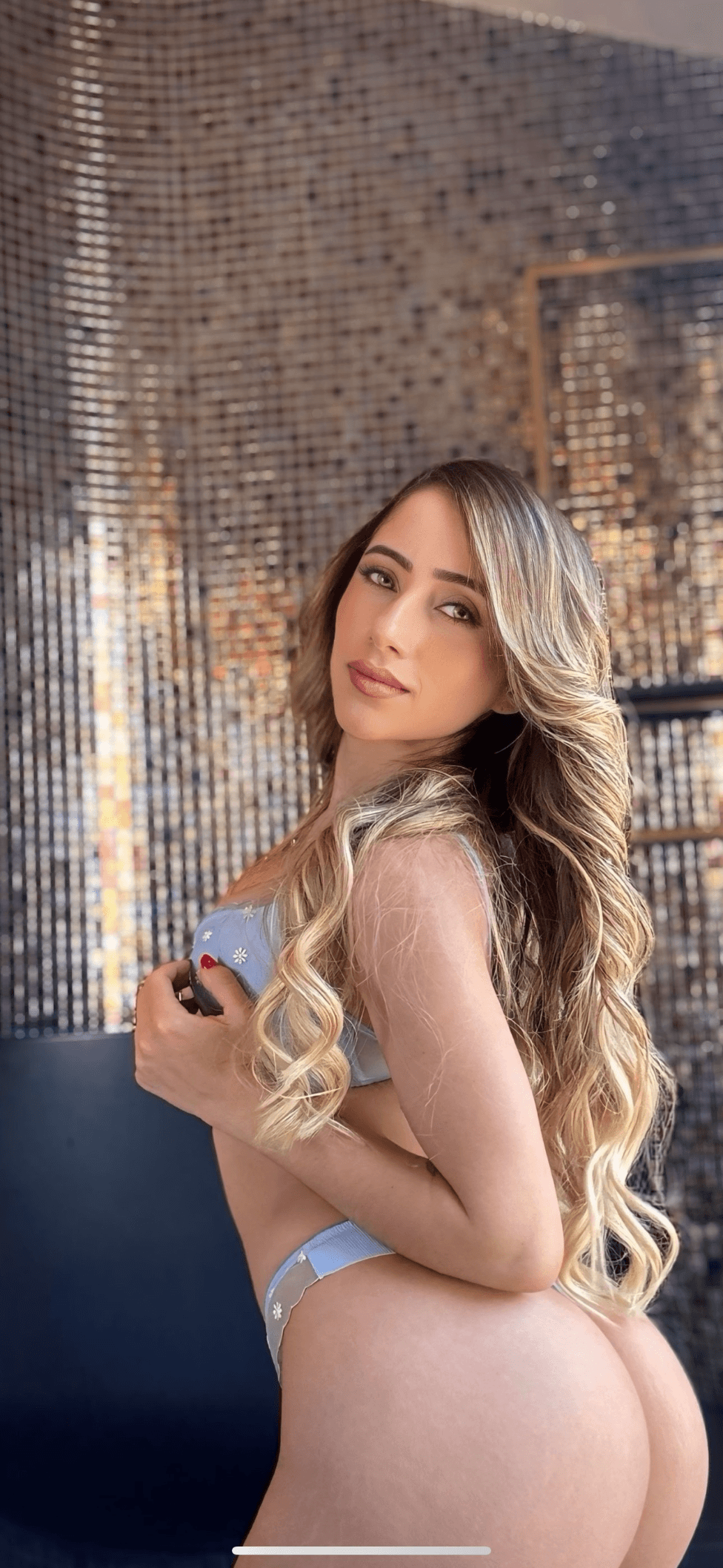 Woman with long wavy hair poses in blue lingerie against a shimmering gold mosaic wall.
