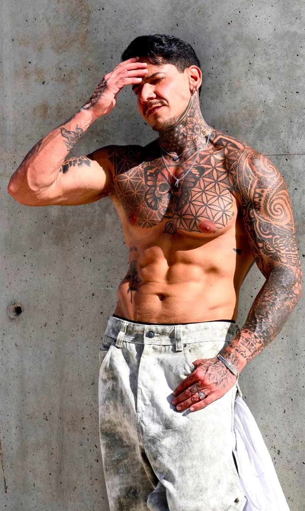 Muscular man with intricate tattoos on his chest and arms posing against a concrete wall.