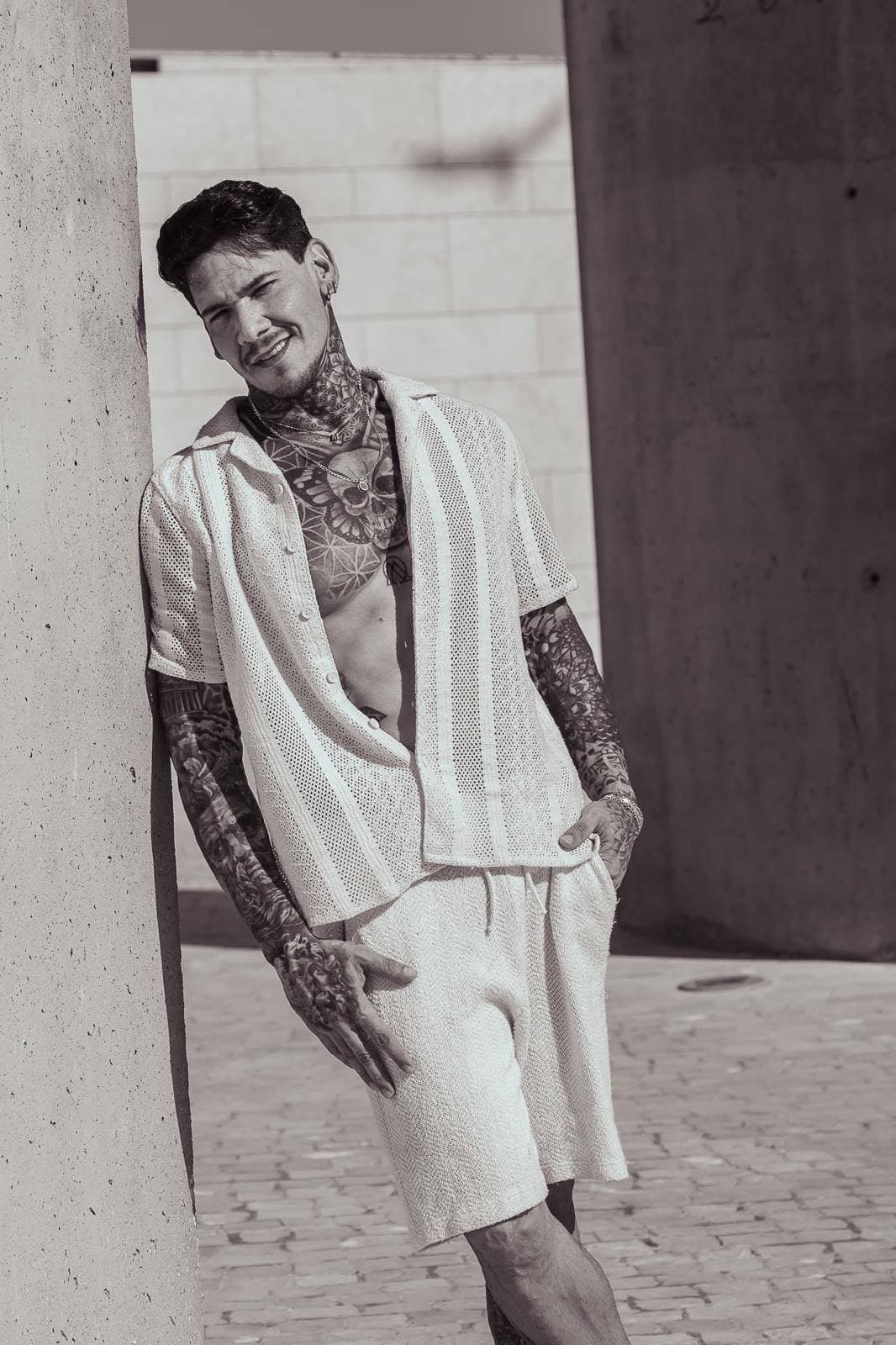 Heavily tattooed man in an open white shirt leaning against a concrete pillar outdoors.